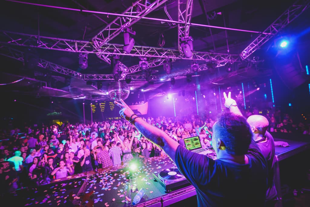 Vista desde la cabina: el DJ saluda a una pista llena bajo luces moradas